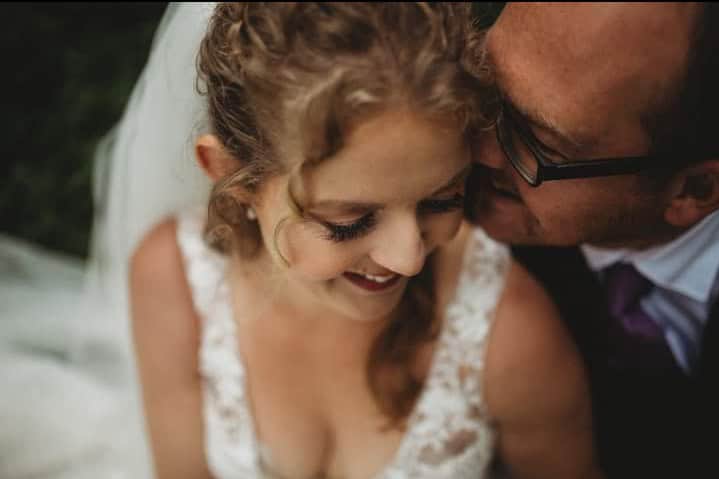 a bride smiles softly with her eyes closed while a groom leans in to kiss her forehead, both framed in a close, intimate setting.